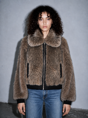 A model with curly dark hair wearing a brown shaggy faux fur jacket with a large collar, black trim, and blue jeans, standing against a gray wall.