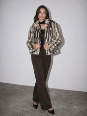 A woman models a cropped faux fur jacket with a beige, brown, and black mottled pattern, worn over a black top and dark brown wide-leg trousers, standing against a plain light gray wall.