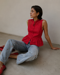 A woman sits on a concrete floor against a plain beige wall, wearing a sleeveless red peplum top with eyelet embroidery, light-wash blue jeans, and red flats.