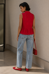 A woman is shown from behind wearing a sleeveless red knit top, light blue jeans with small embroidered details, and red flats, holding a red handbag in a minimalist indoor setting.