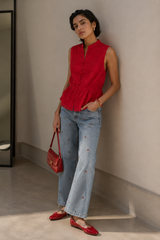 A woman stands against a neutral wall wearing a sleeveless red top, light blue jeans with small embroidered details, red flats, and holding a red handbag.