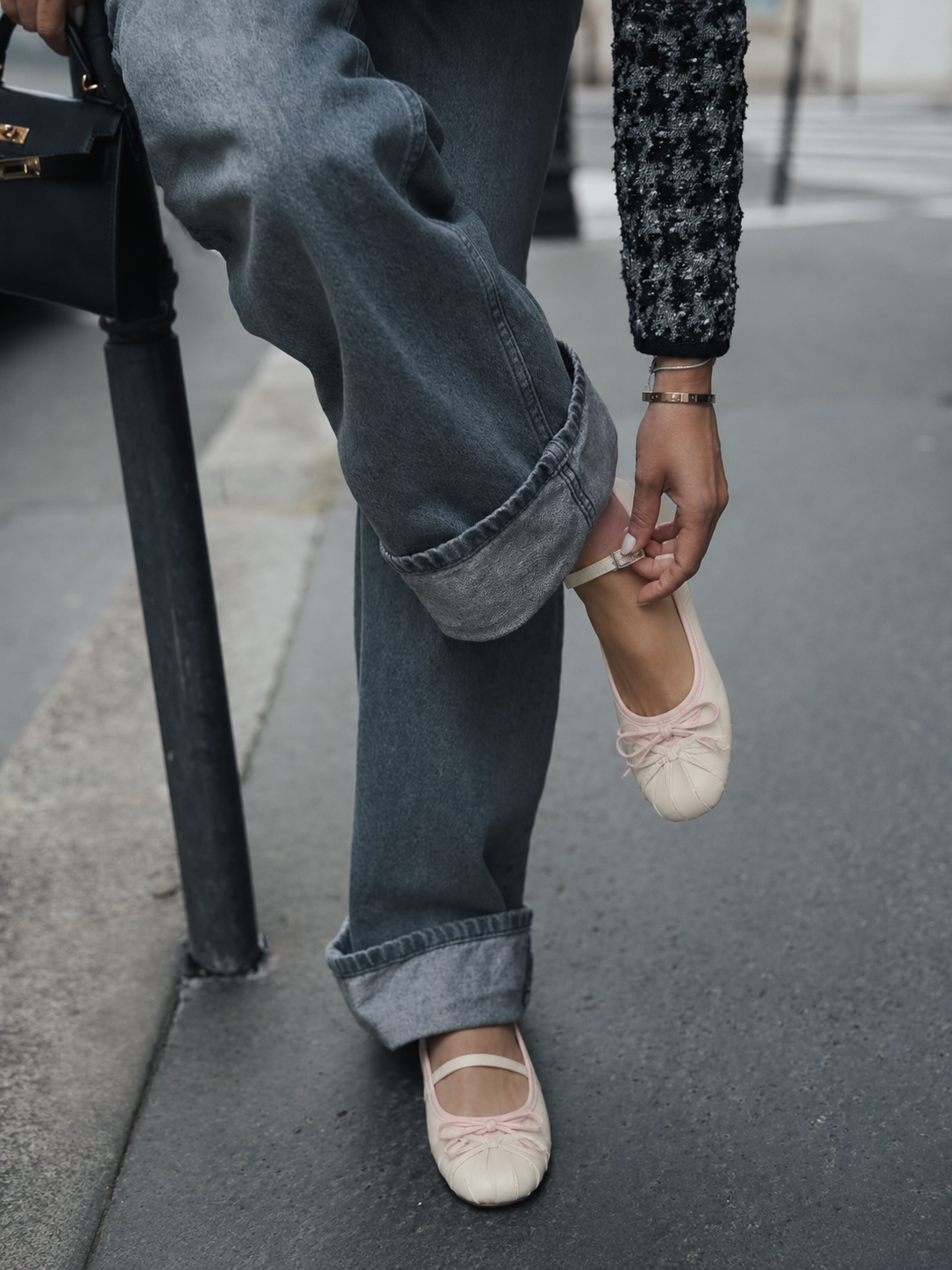A person outdoors on a street or sidewalk wearing loose gray jeans and light pink ballet flats, with one hand adjusting the strap of the shoe. A black handbag and dark textured sleeve are partially visible.