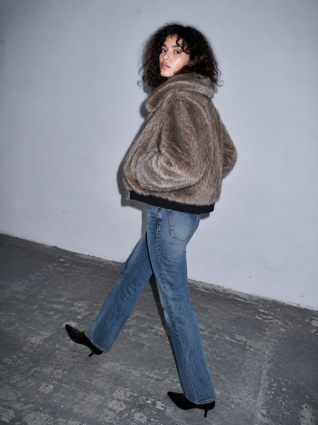 A model wearing a brown fuzzy cropped jacket, blue jeans, and black pointed shoes walks against a plain gray wall and concrete floor.