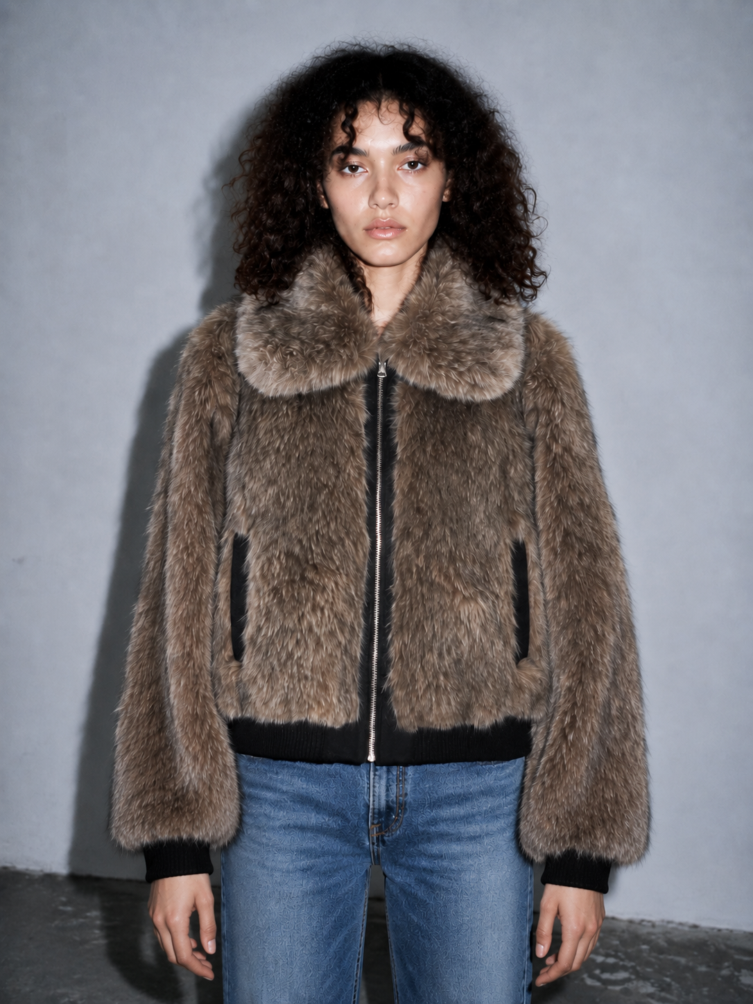 A model with curly dark hair wearing a brown shaggy faux fur jacket with a large collar, black trim, and blue jeans, standing against a gray wall.