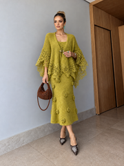 A woman models a matching olive green knit poncho and midi dress set with floral appliqué, holding a brown woven handbag in an indoor setting.