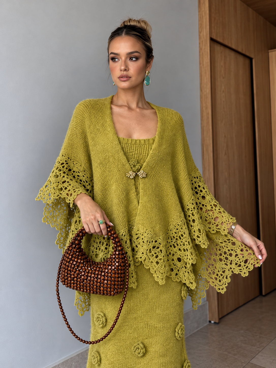 A woman models a matching olive green knit poncho and long-sleeve dress set, holding a brown beaded handbag. The setting is indoors with a gray wall and wooden door in the background.