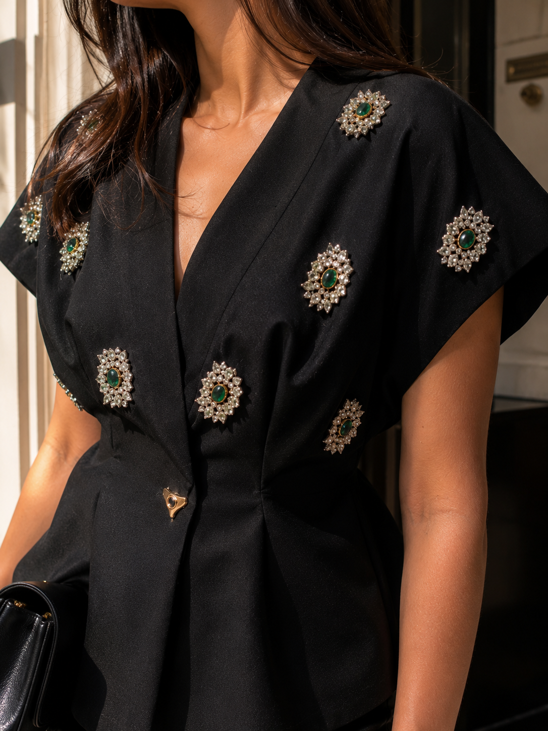A close-up of a woman wearing a black short-sleeve wrap-style top or dress with multiple ornate brooch embellishments featuring green stones and silver-toned settings.