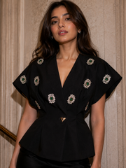 Luxury black peplum top with emerald crystal embellishments, deep V-neckline, structured silhouette, and short sleeves for evening wear.