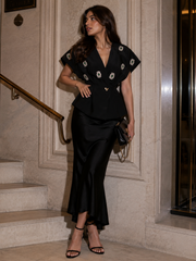 A woman models a black two-piece outfit consisting of a short-sleeve peplum top with white and gold embroidered details and a matching black satin skirt, standing on stone steps.