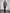 A woman models a cropped faux fur jacket with a beige, brown, and black mottled pattern, worn over a black top and dark brown wide-leg trousers, standing against a plain light gray wall.