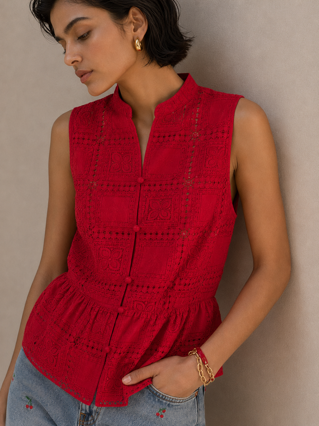 A woman wearing a sleeveless red eyelet blouse with a stand collar and button front, paired with light blue jeans, standing against a neutral beige wall.
